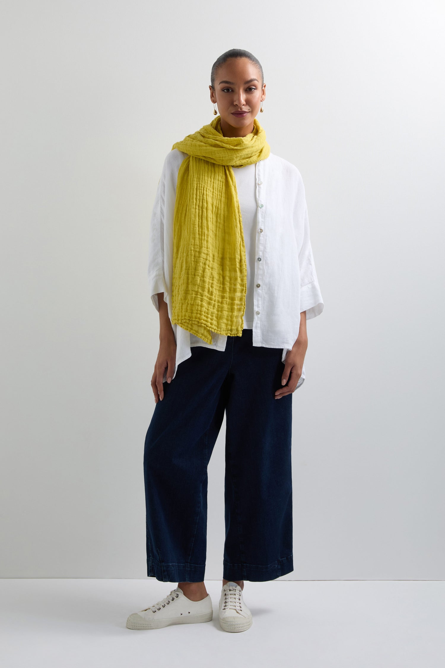 A person in a white shirt, dark blue wide-leg pants, and white sneakers stands against a plain background, adding effortless style with a yellow Crinkle Linen Scarf.