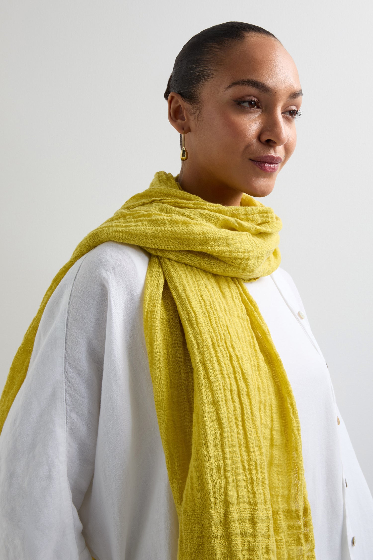 A person in a white shirt wears a Crinkle Linen Scarf with its distinctive yellow, textured design, looking sideways against a plain background.