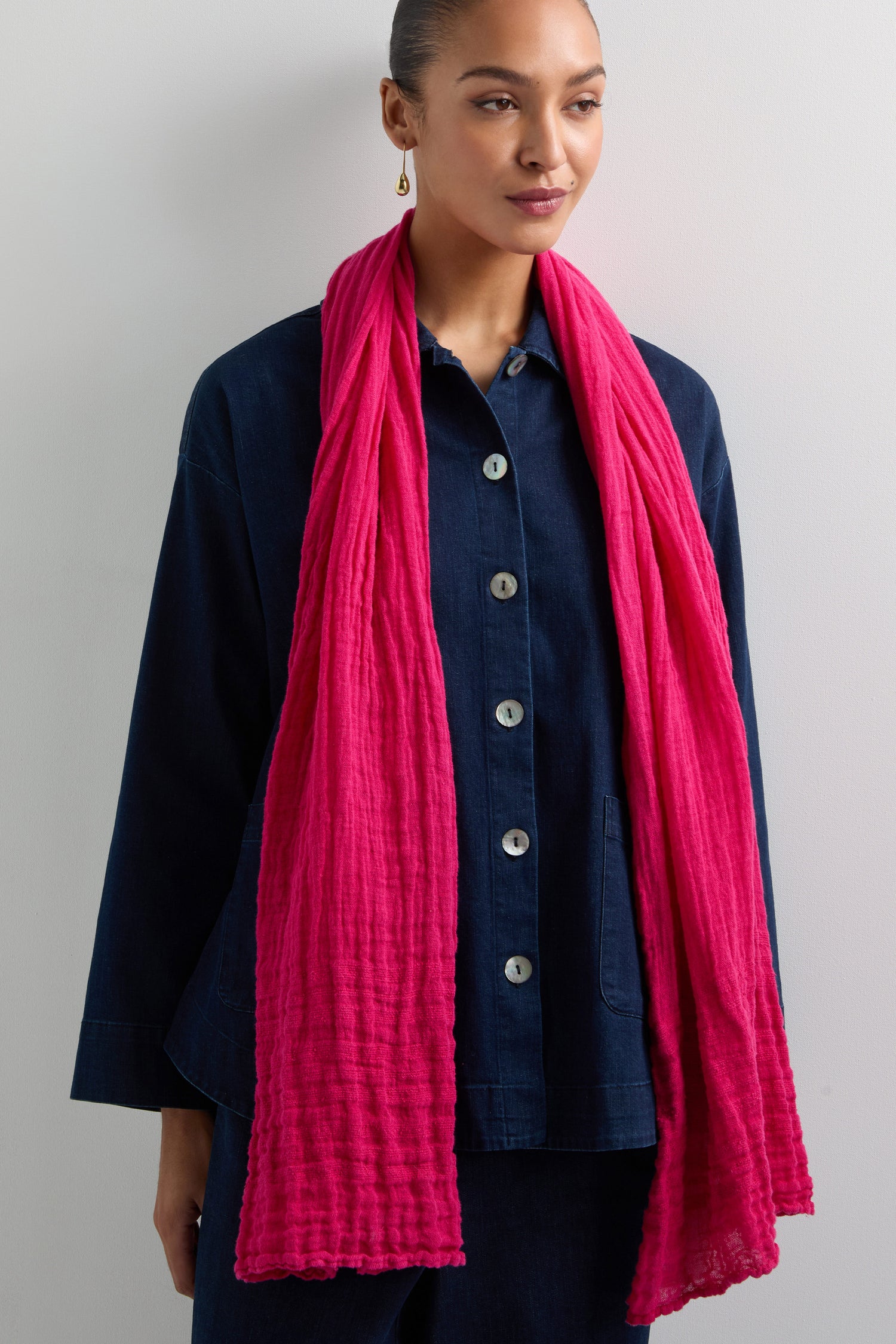 A person in a denim jacket and pants stands against a white background, accessorized with the Crinkle Linen Scarf in bright pink.