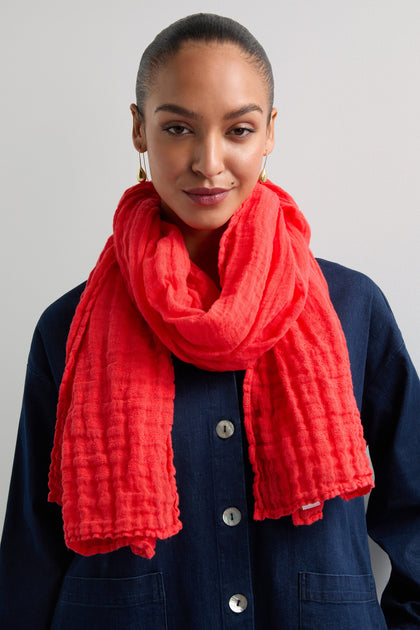 In front of a neutral background, someone sports a dark buttoned shirt and the Crinkle Linen Scarf in bright red. Their hair is neatly pulled back, highlighting the scarf as a vibrant and stylish lightweight accessory.