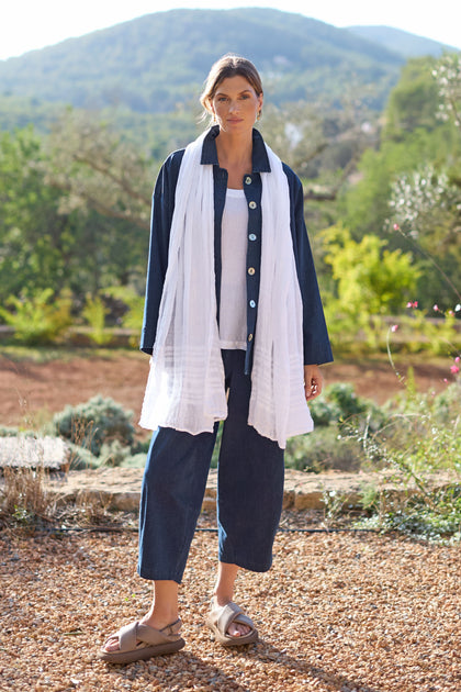 A woman stands on a pebble path, wearing denim and a Crinkle Linen Scarf, with green hills in the background.