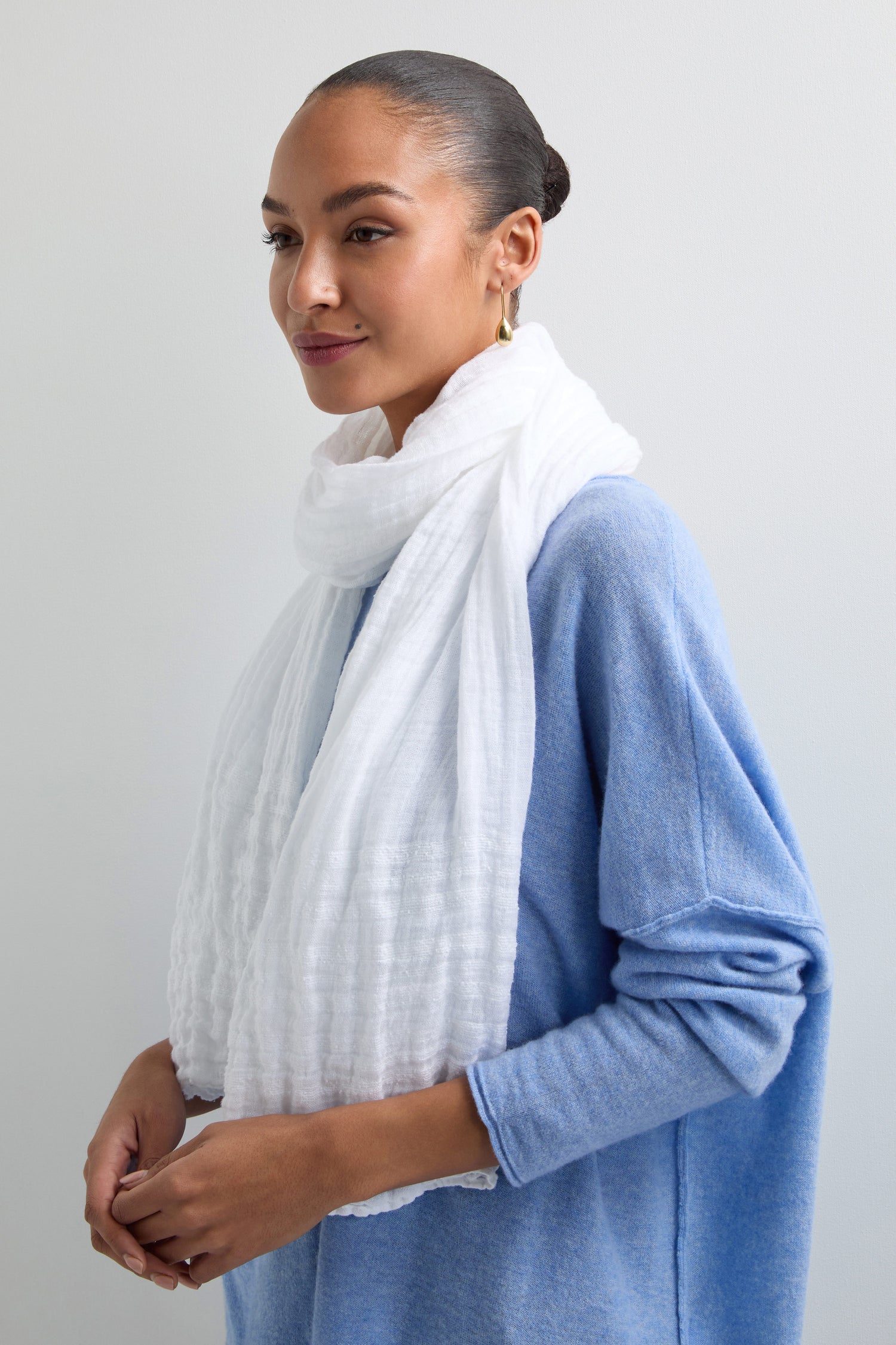 A person in a Crinkle Linen Scarf and light blue sweater, hoop earrings, and hair tied back, stands against a light background.