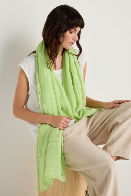 A woman sits on a wooden stool, wearing a sleeveless white top, beige pants, and the Crinkle Linen Scarf in light green, looking down with one arm resting on her knee.