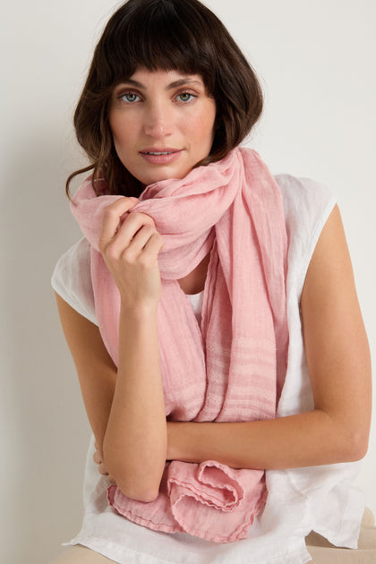 Woman with wavy brown hair wearing a white top and the Crinkle Linen Scarf in light pink, posed against a plain light background.