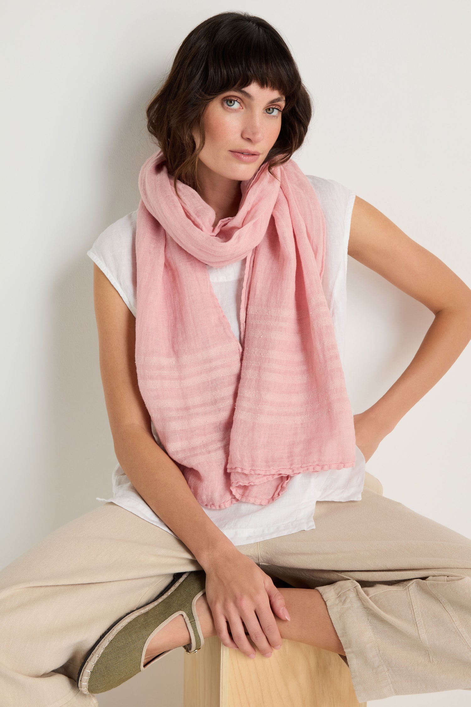 A woman sits on a wooden stool against a plain white background, wearing a Crinkle Linen Scarf in light pink, paired with a white top, beige pants, and olive green shoes.