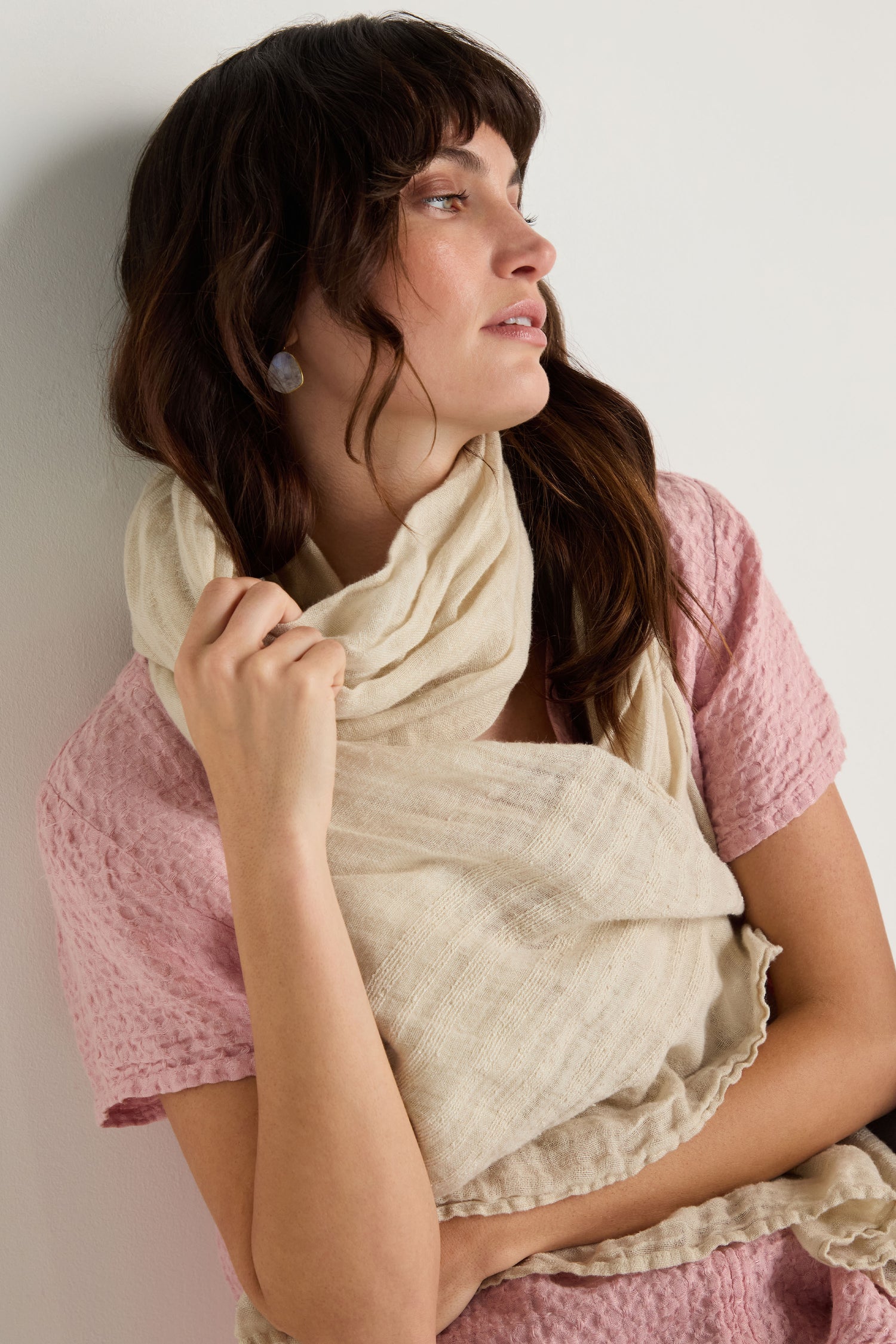 A woman leans against a light wall, looking to the side, wearing a textured pink top paired with the Crinkle Linen Scarf.