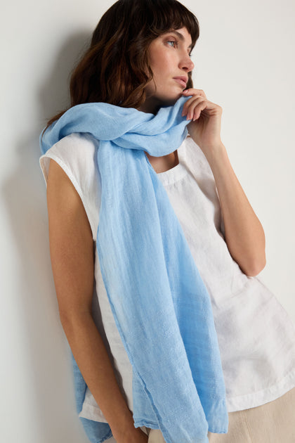 A woman stands against a plain white wall, wearing a loose white top and the Crinkle Linen Scarf in light blue, looking to the side with her hand gently touching her face.