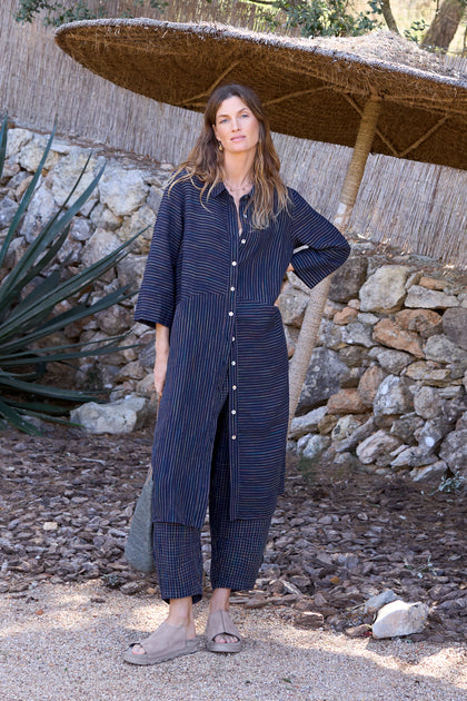 A woman in a Navy Space Dye Stripe Shirt Linen Dress and sandals stands outdoors on a gravel path, with a straw umbrella and stone wall in the background.