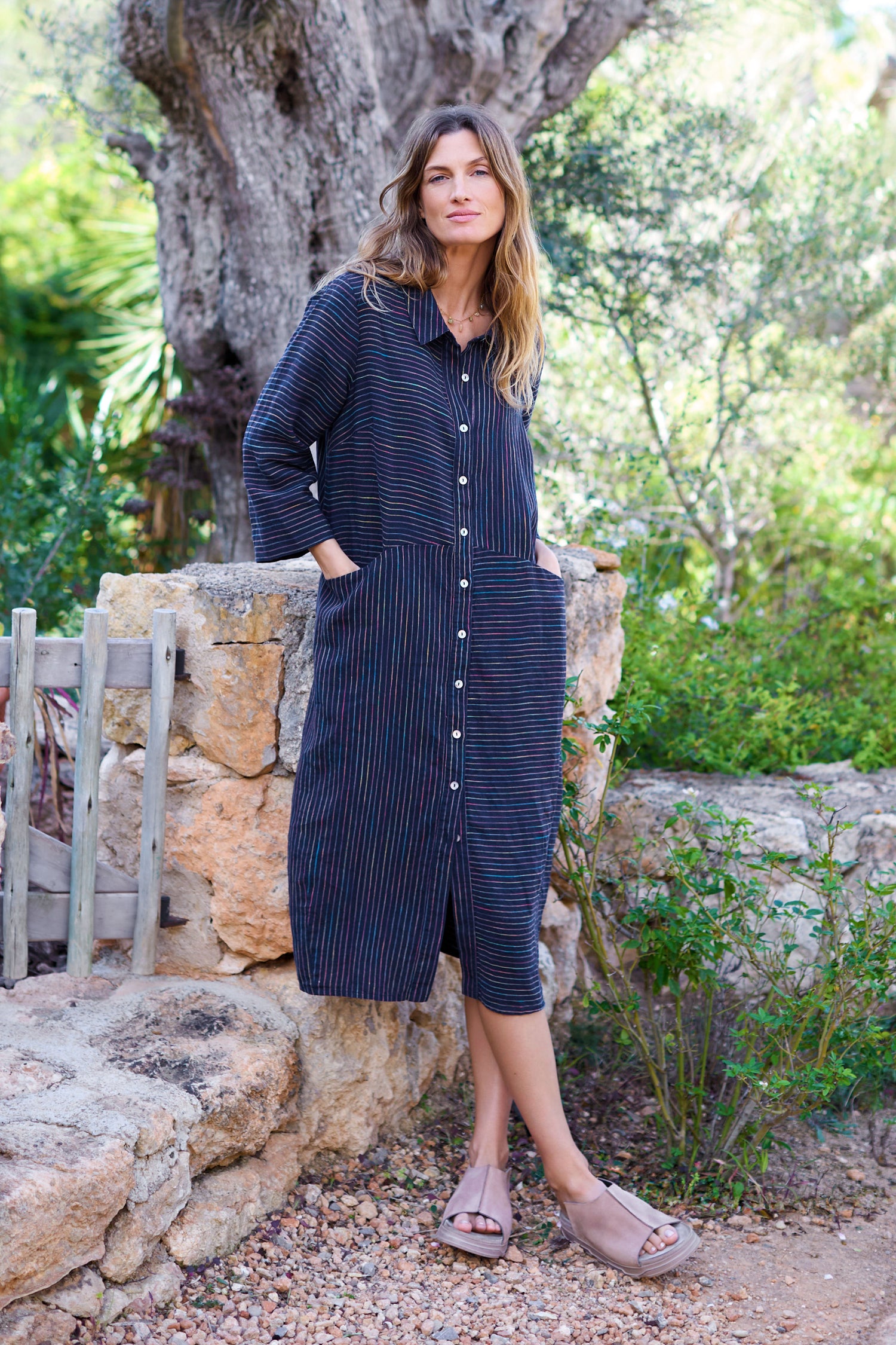 A woman in a classic silhouette stands against a stone wall, wearing the Linen Navy Space Dye Stripe Shirt Dress, with one hand in her pocket while embracing the garden setting.