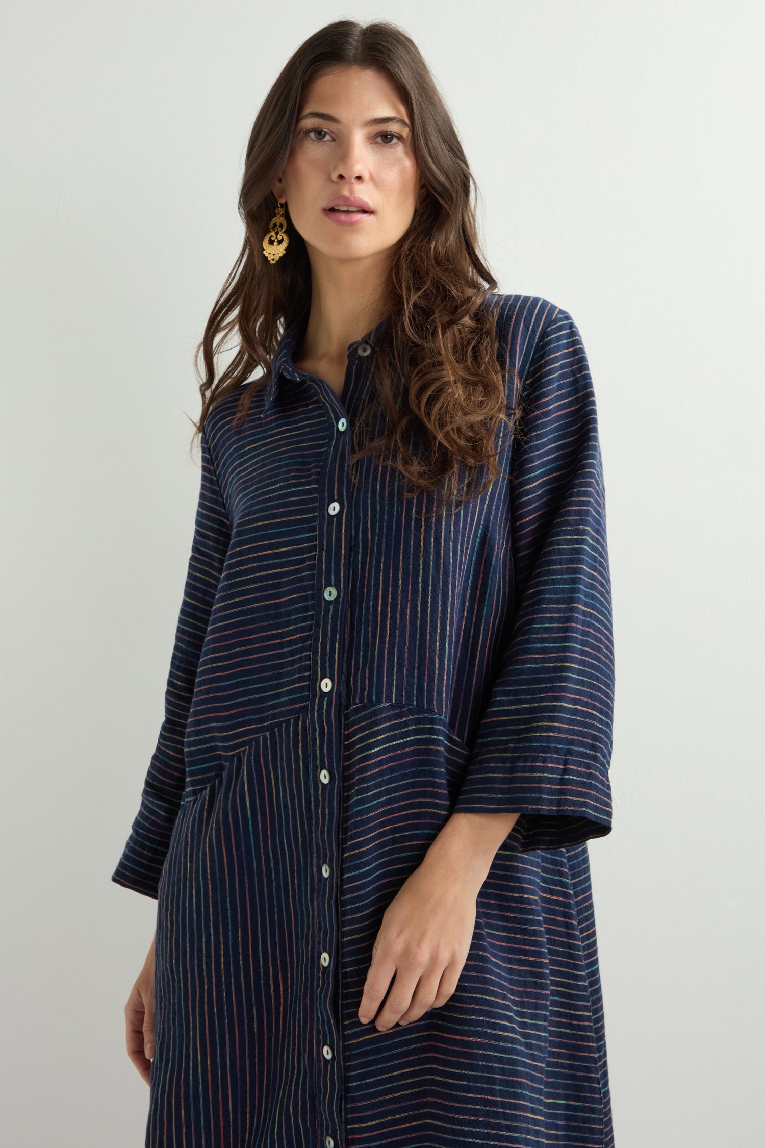 A person wearing a Linen Navy Space Dye Stripe Shirt Dress with a button-up front and gold earrings stands against a plain white background.