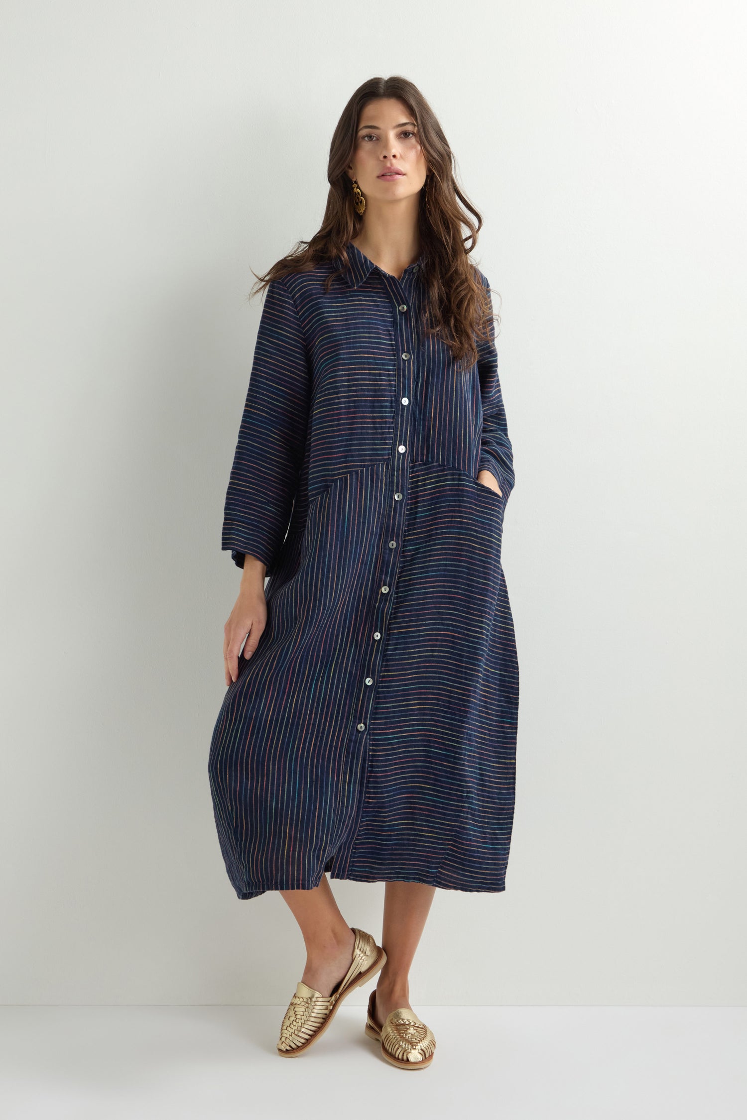 A woman stands against a white background, elegantly wearing a Linen Navy Space Dye Stripe Shirt Dress with gold shoes.