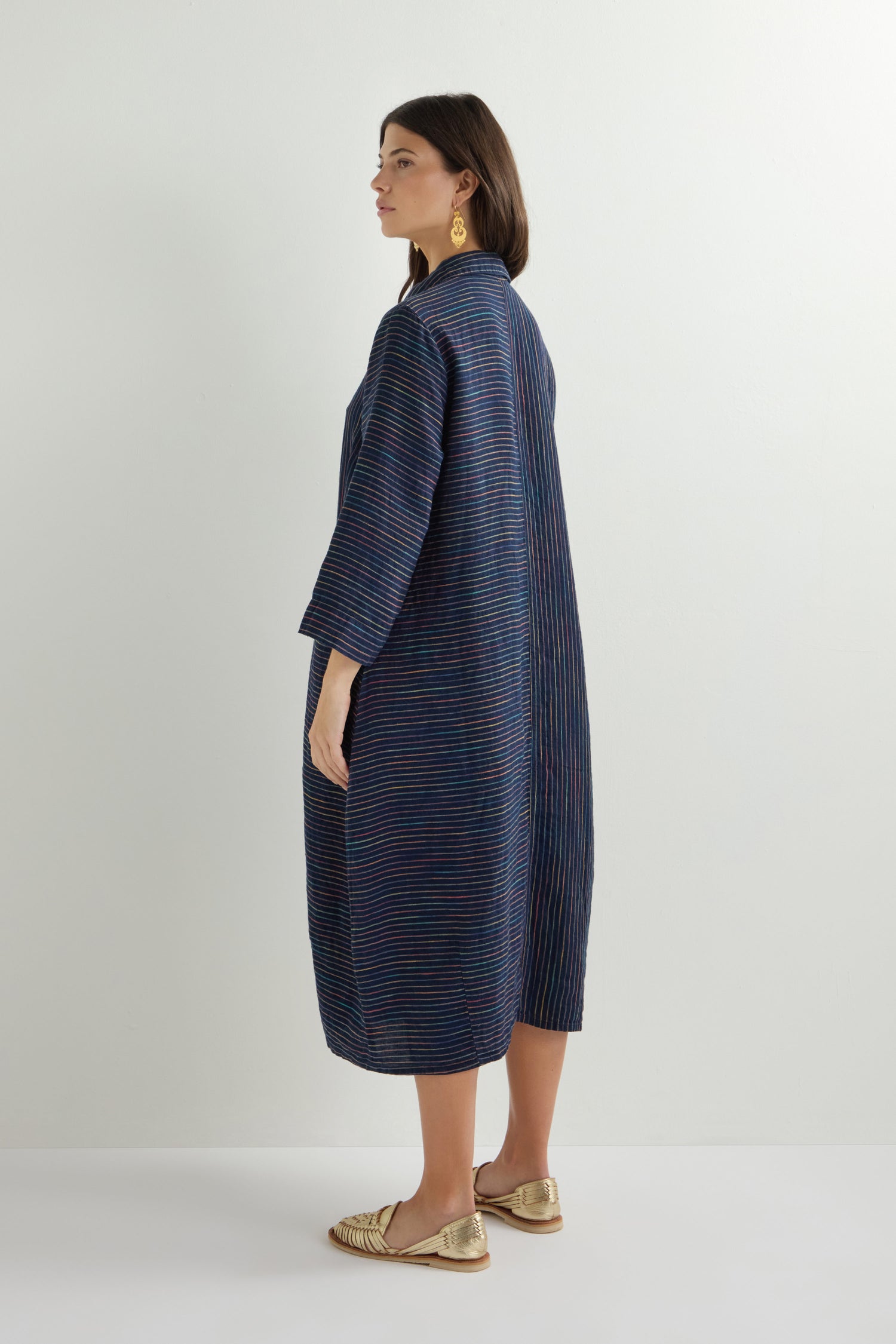 A woman stands sideways in a navy space dye stripe shirt dress, with shoulder-length hair that complements her gold sandals and large gold earrings against a plain white background. The dress is the Linen Navy Space Dye Stripe Shirt Dress.