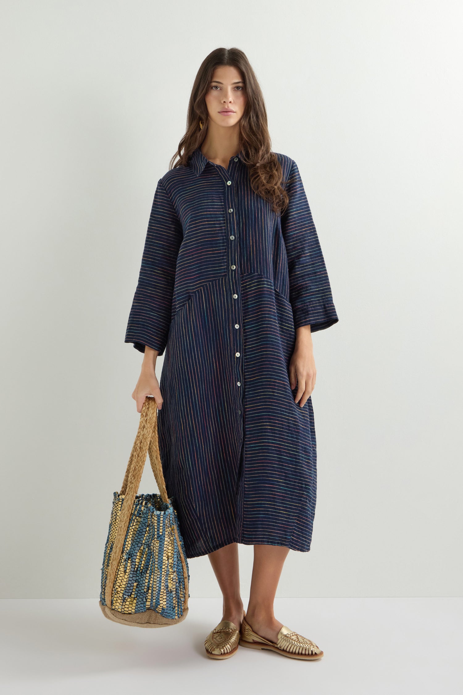 A woman in a Linen Navy Space Dye Stripe Shirt Dress holds a woven bag and stands wearing gold slip-on shoes.