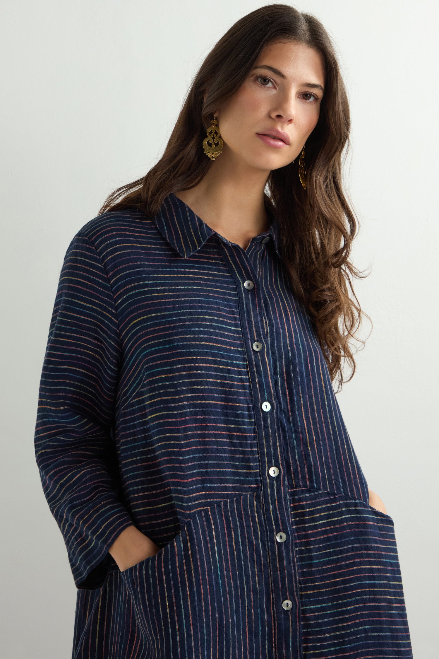 A person with long hair, wearing gold earrings and a Linen Navy Space Dye Stripe Shirt Dress featuring pockets, stands against a plain background.