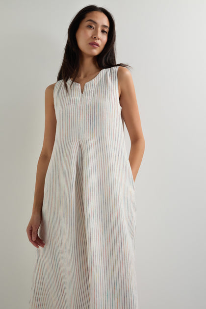 A woman wears the White Space Dye Stripe Sleeveless Linen Dress, featuring an A-line silhouette with vertical pastel stripes, while standing against a plain light background.