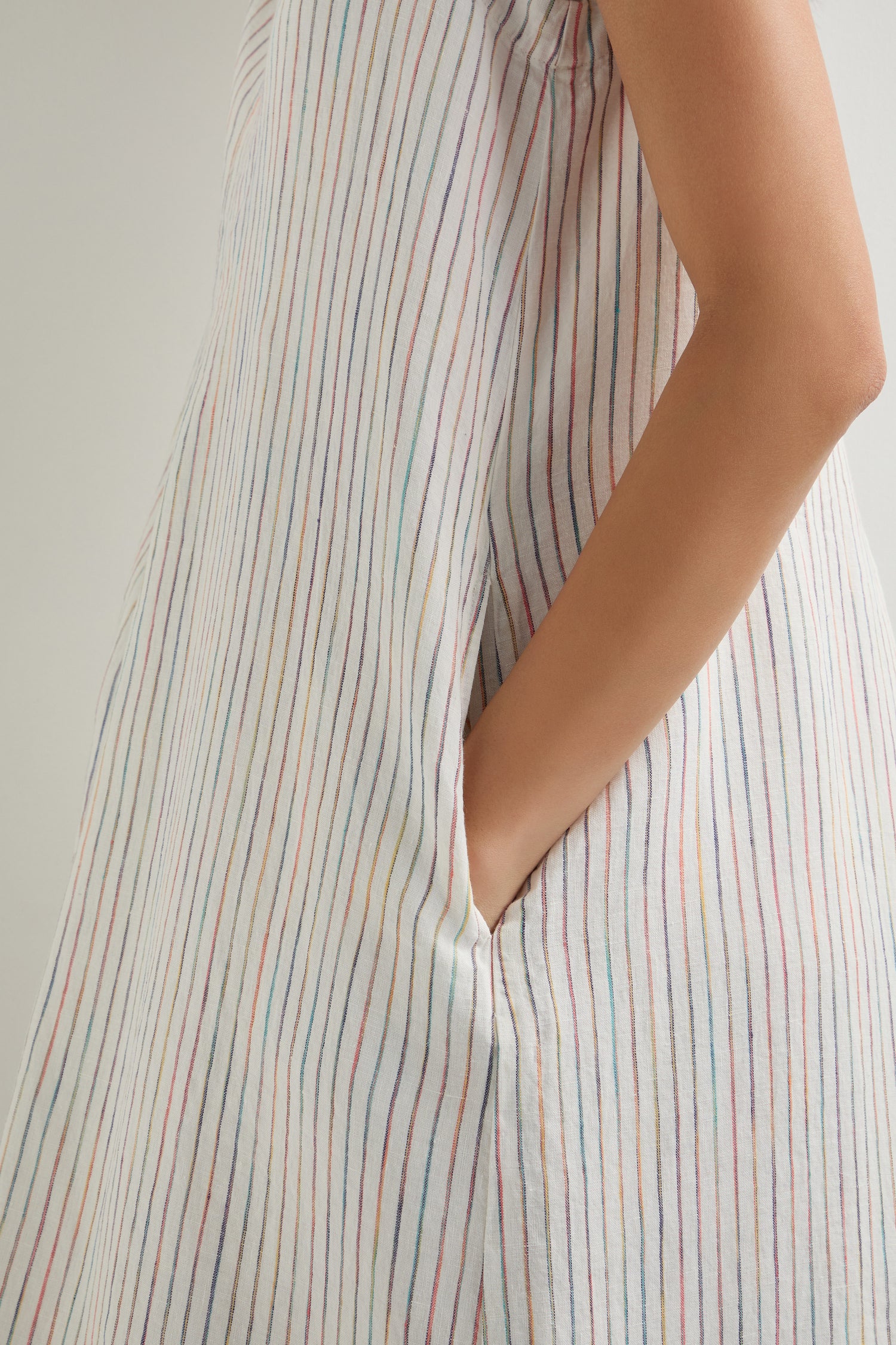 A person wearing the White Space Dye Stripe Sleeveless Linen Dress, featuring thin multicolored vertical stripes, an A-line silhouette, and side pockets, shown from the side with one hand in a pocket.