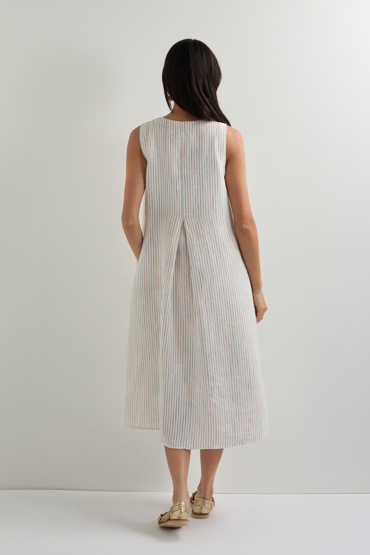 A woman faces away, wearing the White Space Dye Stripe Sleeveless Linen Dress—an A-line dress with subtle stripes and back pleats—paired with flat sandals against a plain light background.
