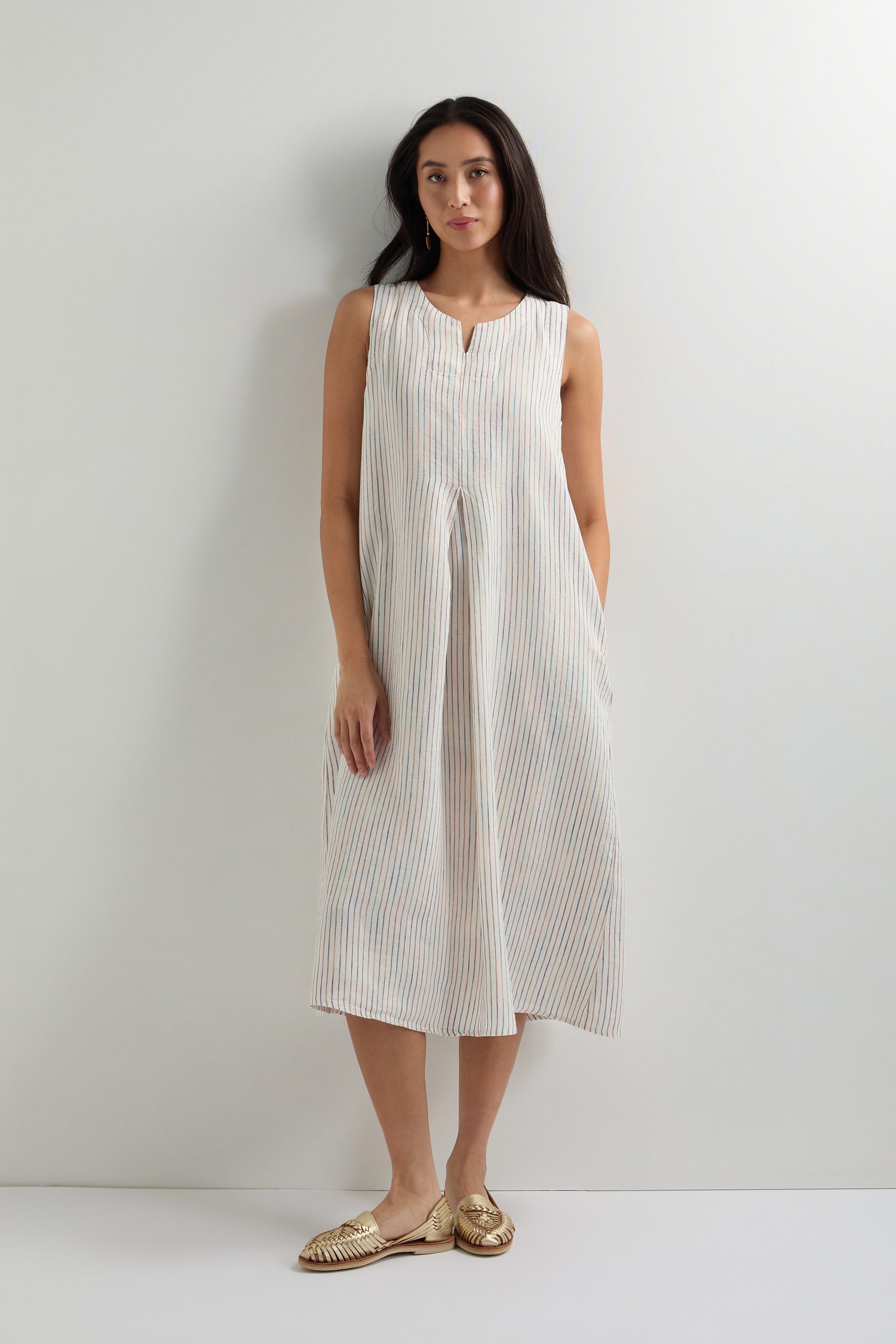 A woman stands against a plain light-colored wall wearing a White Space Dye Stripe Sleeveless Linen Dress with an A-line silhouette and woven gold slip-on shoes.