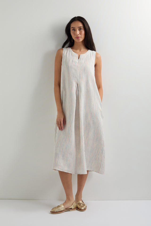 A woman stands against a plain light-colored wall wearing a White Space Dye Stripe Sleeveless Linen Dress with an A-line silhouette and woven gold slip-on shoes.