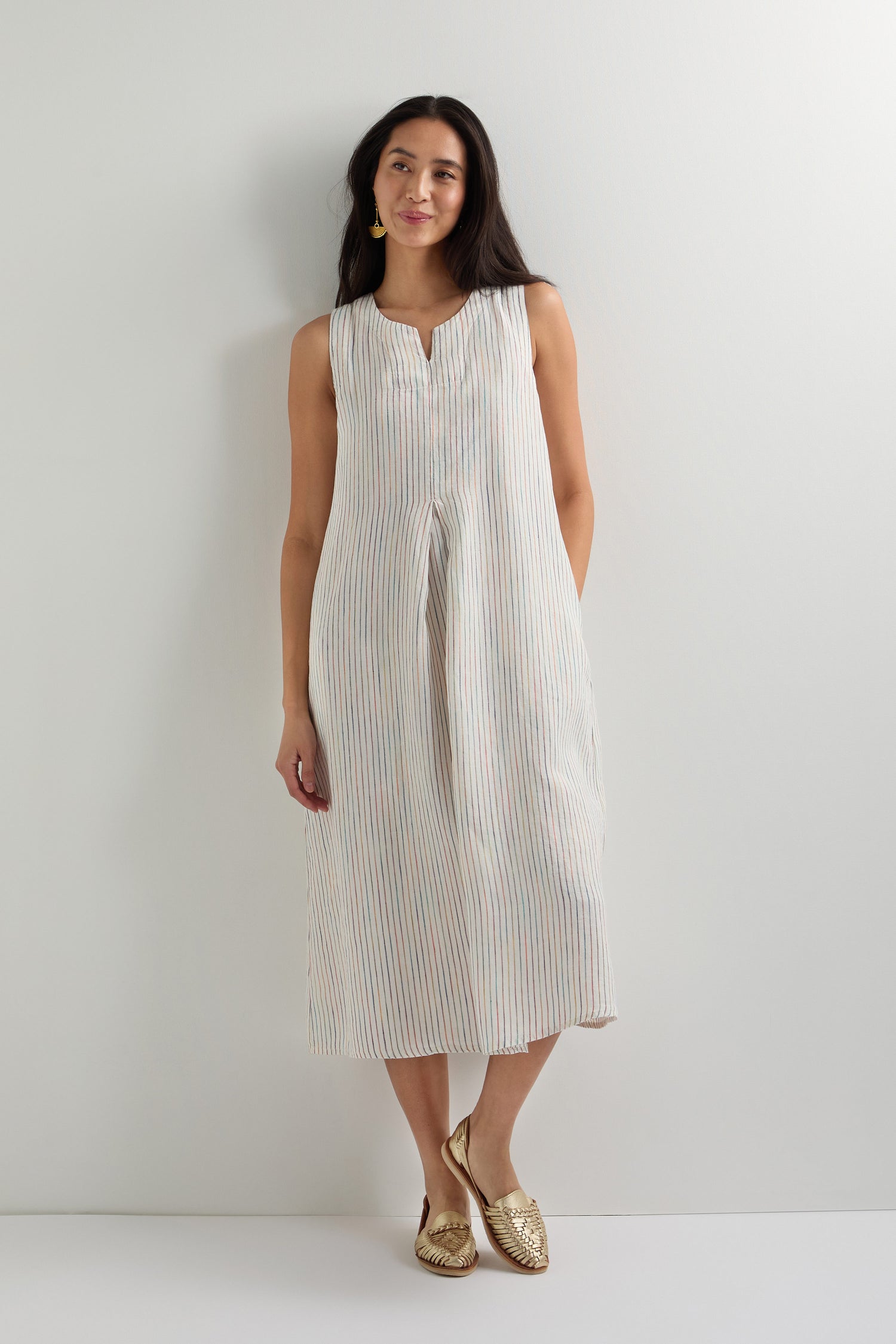 A woman stands against a white wall wearing the White Space Dye Stripe Sleeveless Linen Dress and gold flats.