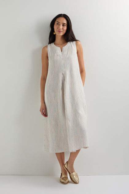 A woman stands against a white wall wearing the White Space Dye Stripe Sleeveless Linen Dress and gold flats.