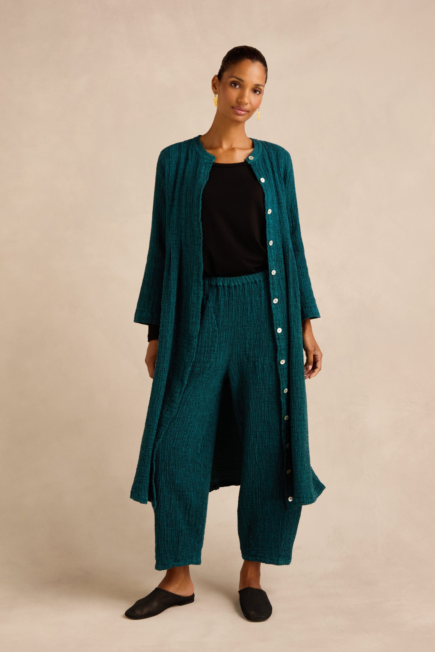 A woman stands wearing a teal Crinkle Double Face Linen Dress styled as a long button-down top, paired with matching pants, a black shirt, and black shoes against a neutral background.