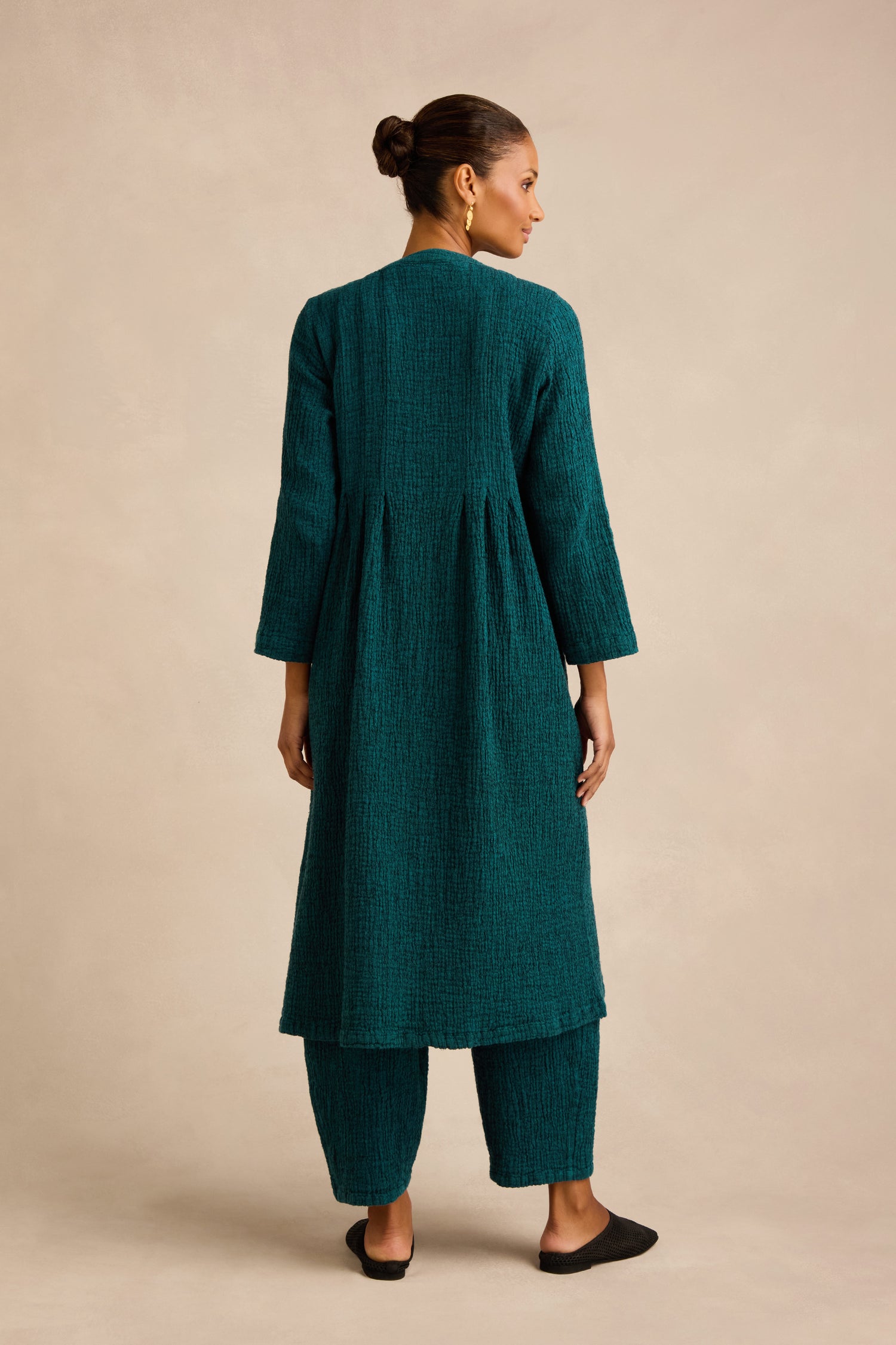 A woman stands facing away, wearing the Crinkle Double Face Linen Dress in teal, styled as a long tunic with matching pants and black shoes, set against a plain beige background.