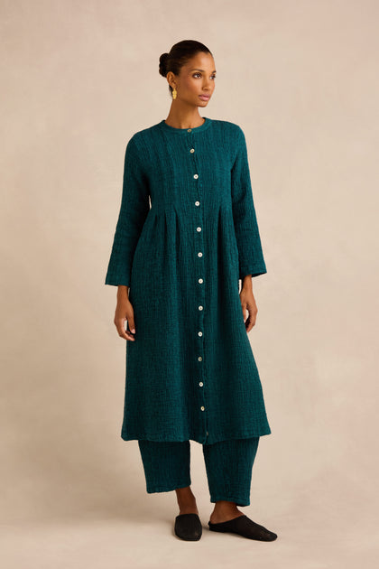 A woman stands against a plain beige background wearing a teal Crinkle Double Face Linen Dress paired with matching wide pants and black shoes.