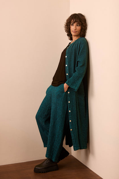 A person with curly hair leans against a beige wall, wearing the Crinkle Linen Blend Shirt Dress in teal, paired with black platform shoes.