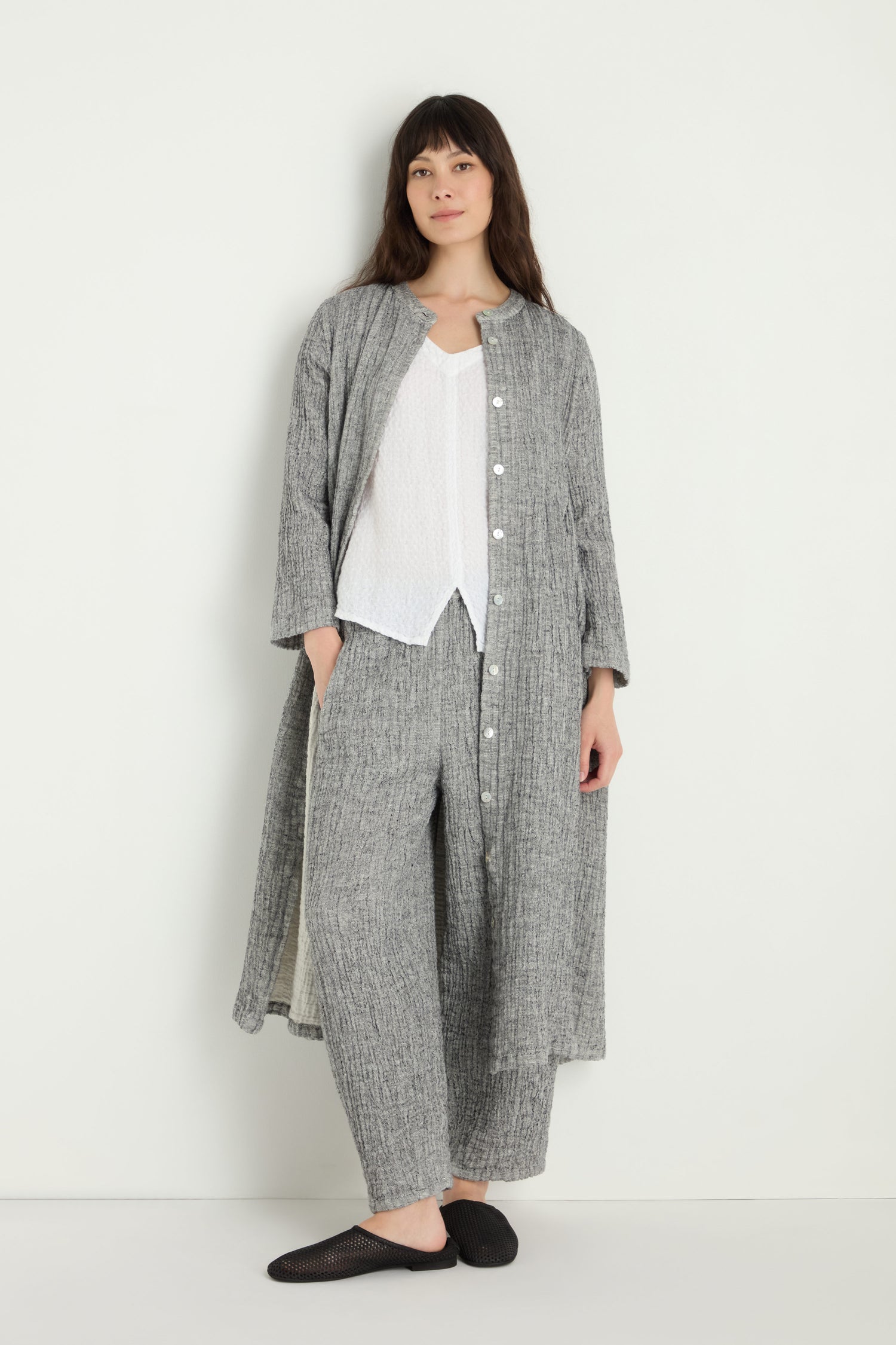 A woman stands against a plain light wall, wearing the Crinkle Double Face Linen Shirt Dress in gray, styled as a coat over a white top and matching pants, with black slip-on shoes.