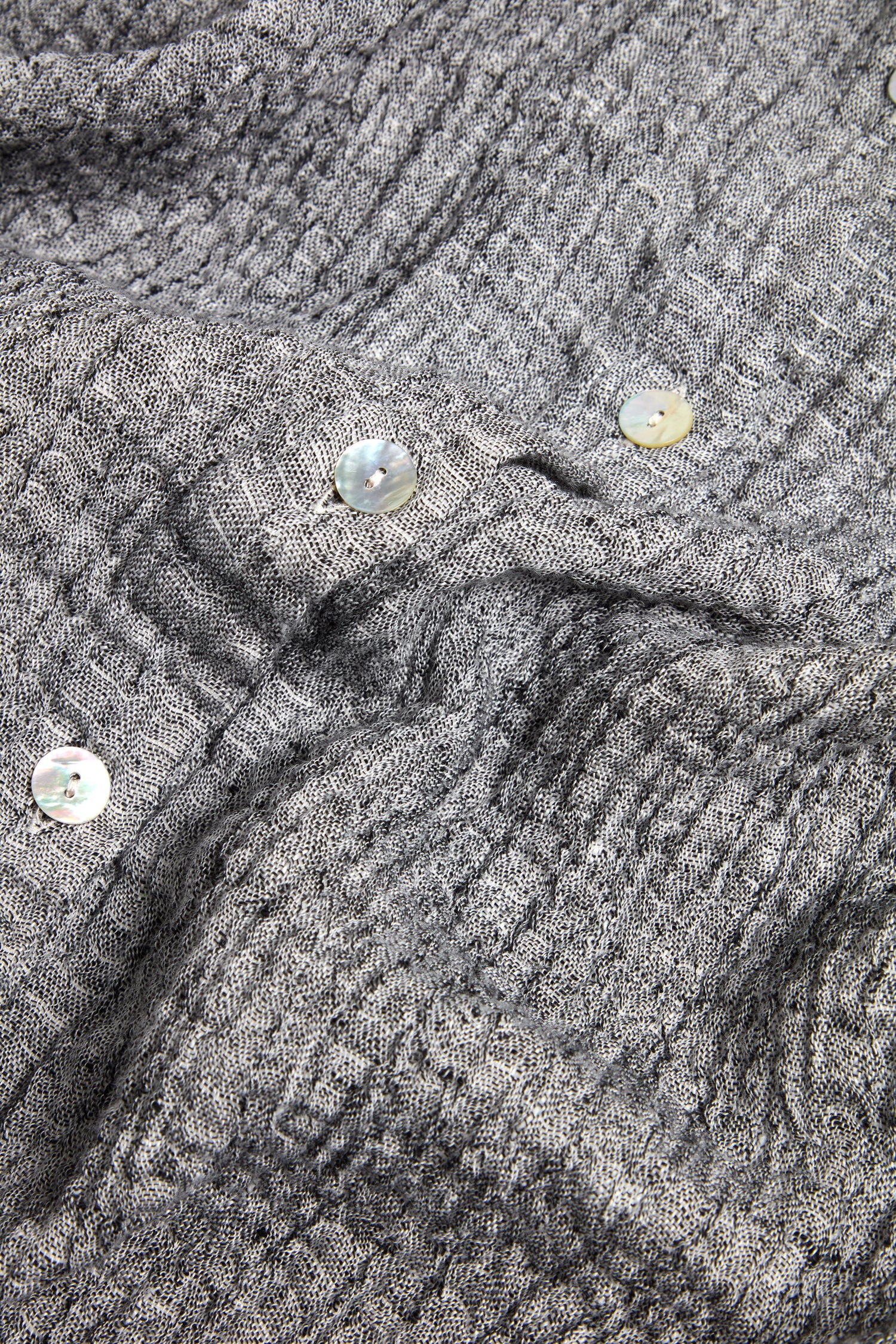 Close-up of Crinkle Double Face Linen Shirt Dress fabric, featuring a textured gray surface with four white buttons scattered on top.
