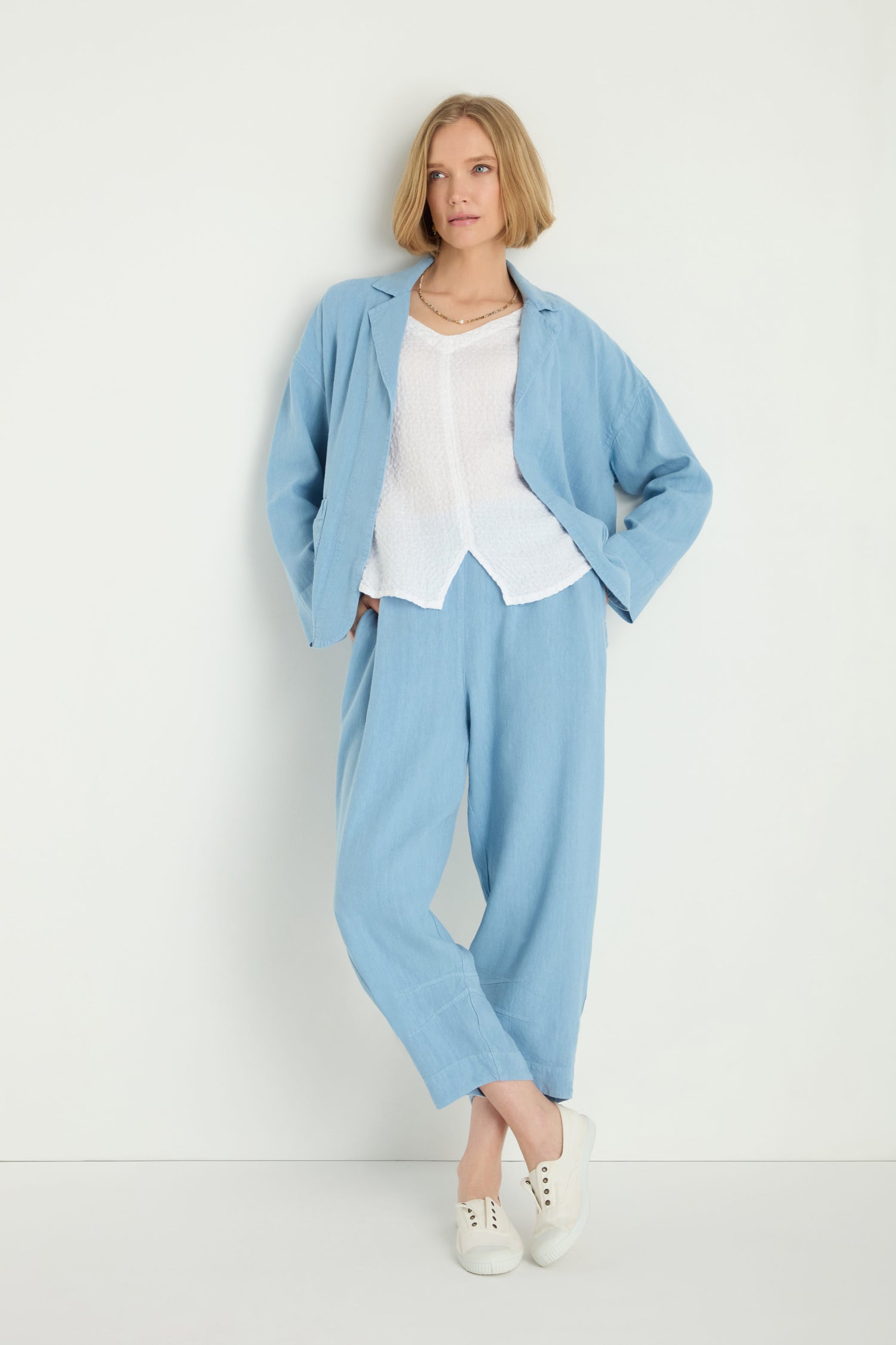 A person stands against a white background wearing the Twisted Linen Boxy Jacket, featuring a light blue hue and relaxed silhouette, paired with a white blouse and white sneakers.