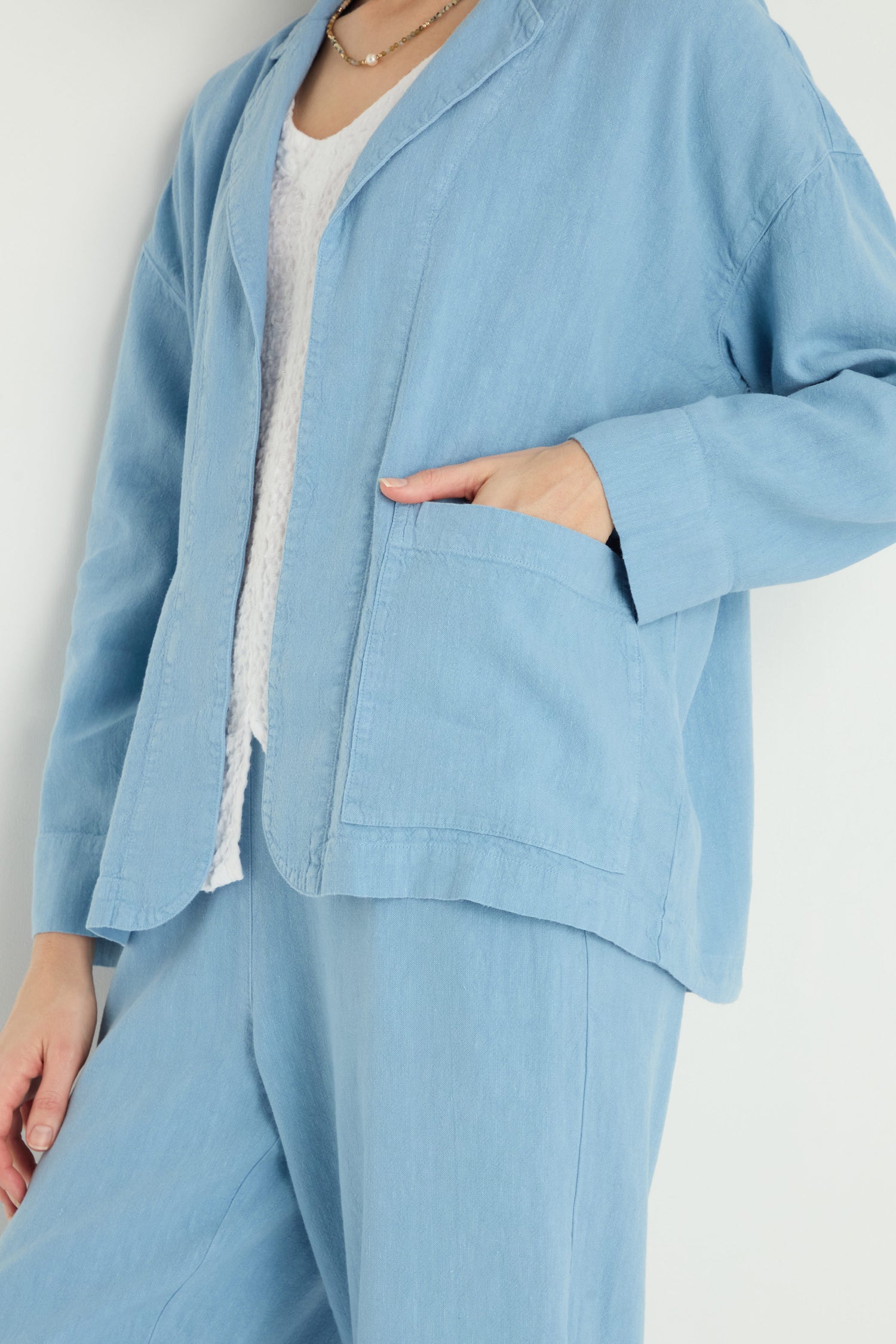 Person wearing the Twisted Linen Boxy Jacket in light blue with matching pants and a white shirt, standing with one hand in the jacket pocket.