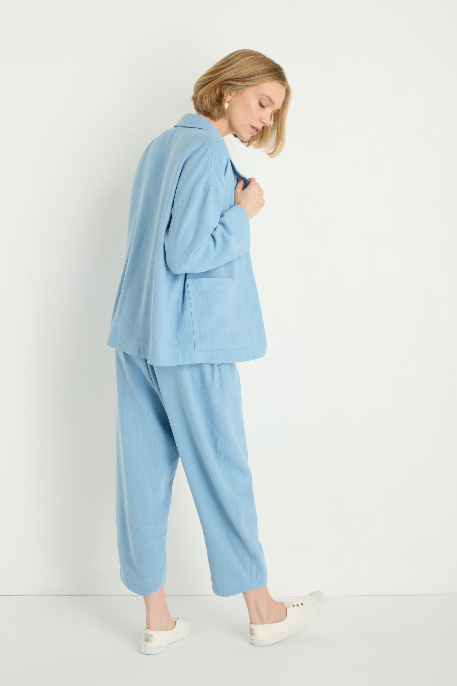 A woman stands sideways against a white background, wearing the Twisted Linen Boxy Jacket in a light blue, relaxed silhouette suit paired with white sneakers.