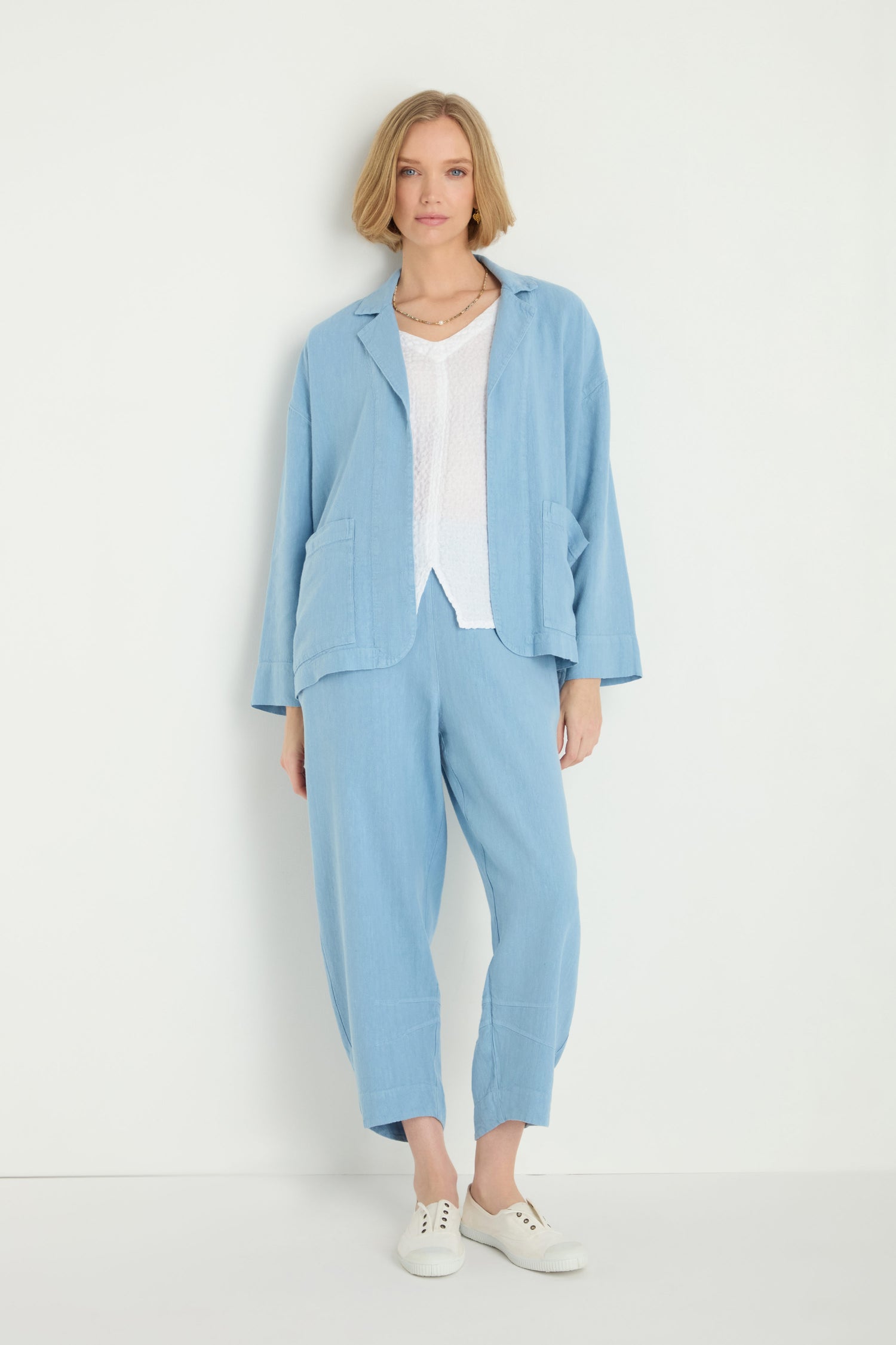 A woman stands against a white wall wearing the Twisted Linen Boxy Jacket, styled with a white top and white sneakers.