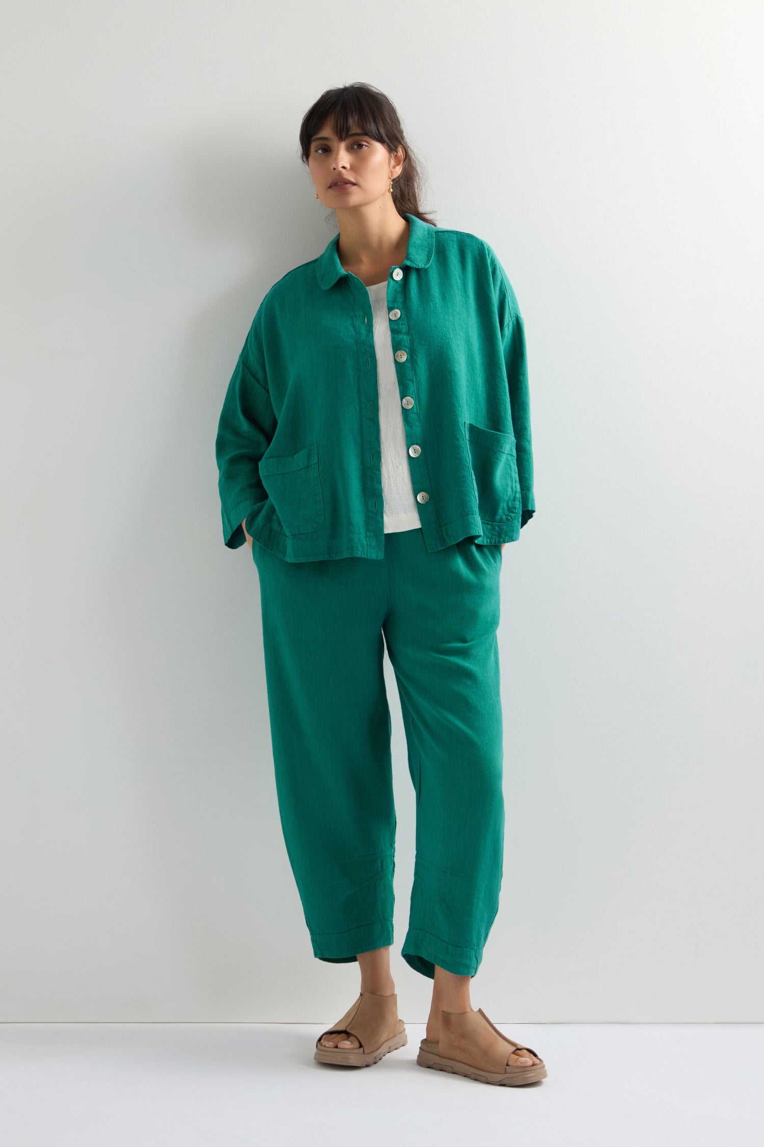 Against a white wall, someone wears the Twisted Linen Boxy Jacket in an oversized green style with matching pants, a crisp white shirt, and sandals.