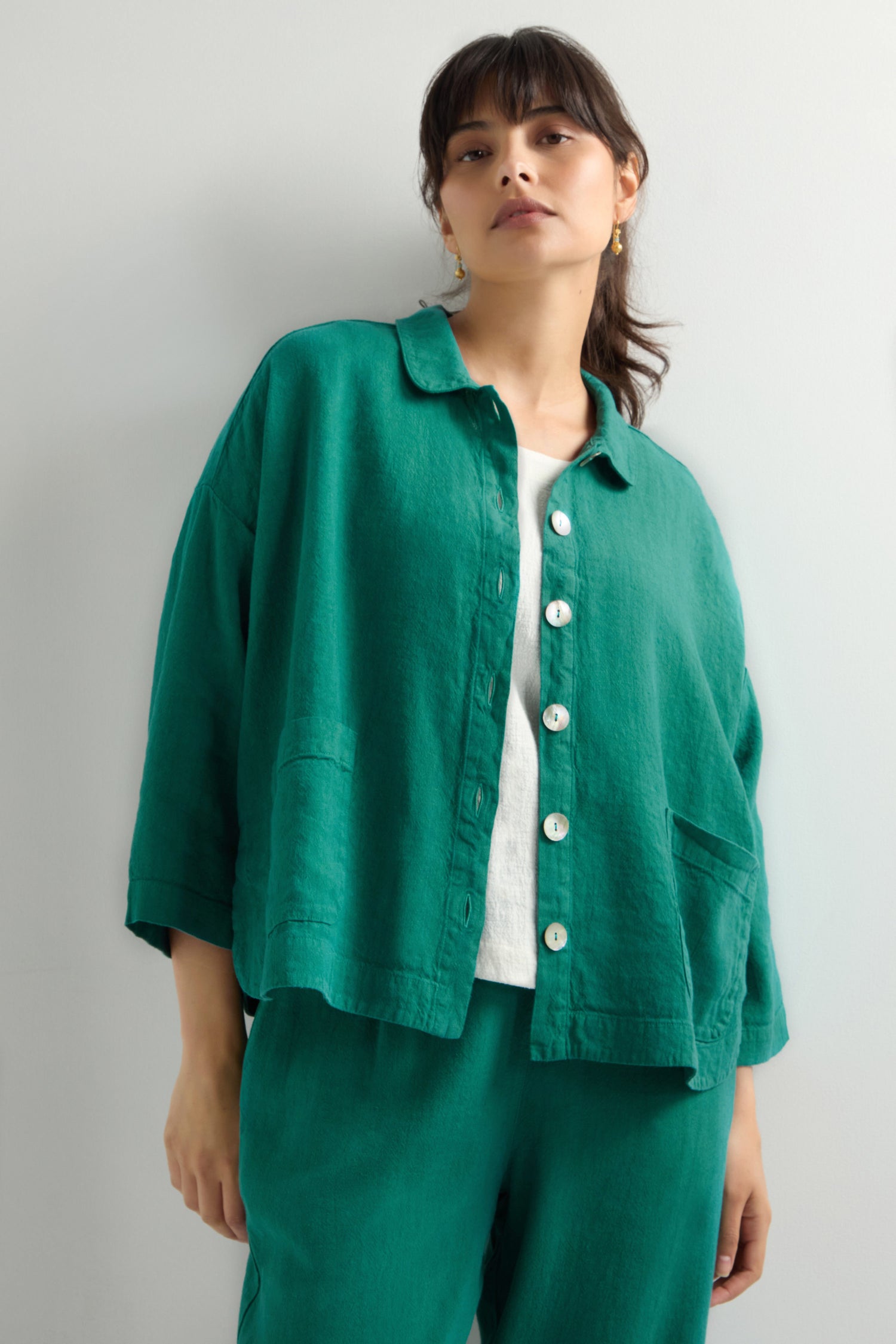 A person wears the Twisted Linen Boxy Jacket, a loose green button-up with an oversized fit over a white top, paired with green breathable linen blend pants, standing against a plain background.