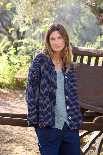 Wearing a plaid shirt and blue pants, a person stands outdoors in front of a wooden structure. They are layered with the Linen Navy Space Dye Check Boxy Jacket, surrounded by trees and greenery.