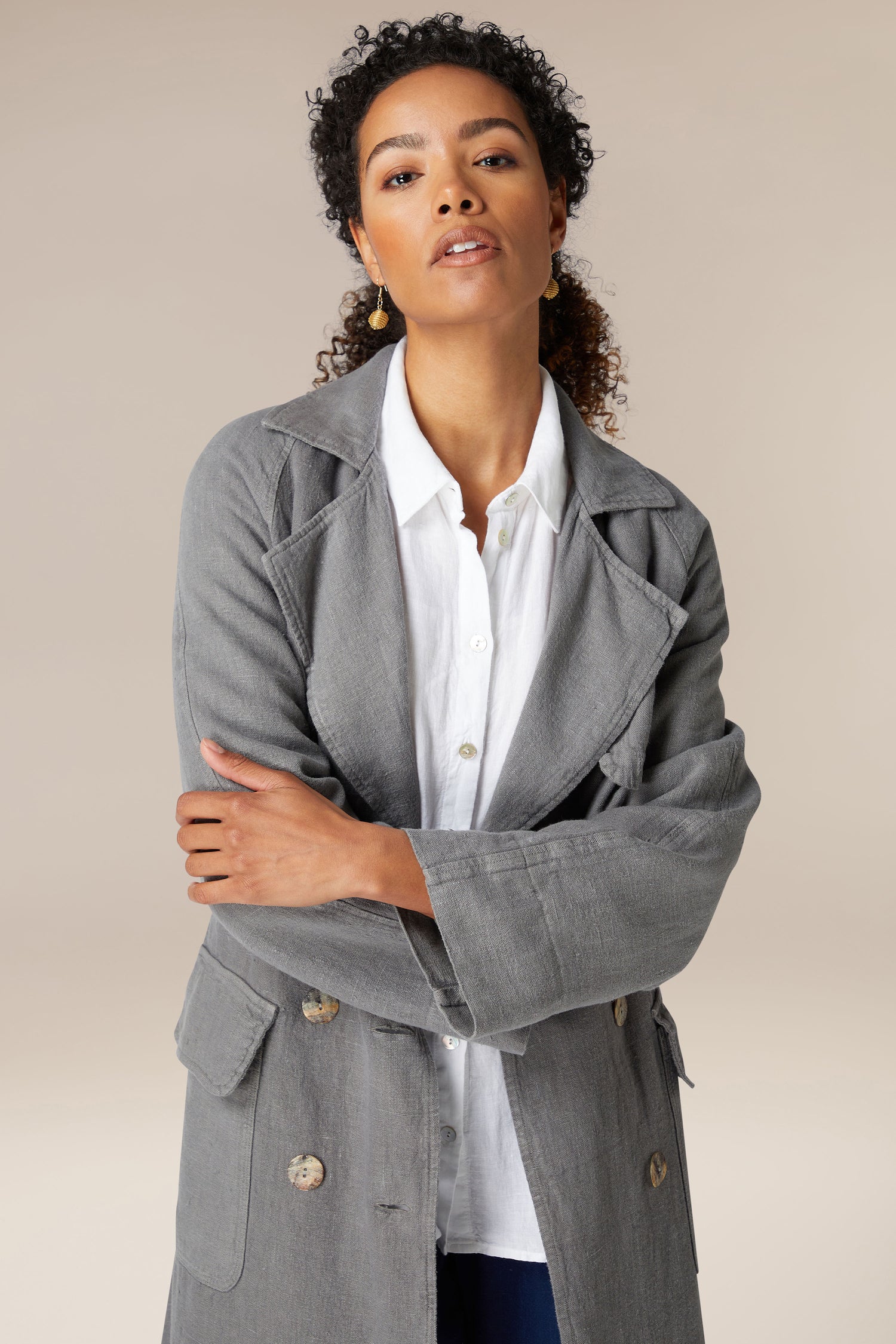 A confident woman with curly hair wearing the Grayson Linen Trench Coat and white shirt, standing with arms crossed against a neutral background.