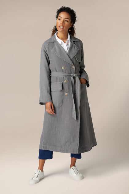 A woman stands against a neutral background, wearing a grey lightweight Linen Trench Coat over a white shirt and navy trousers, complemented by white sneakers.