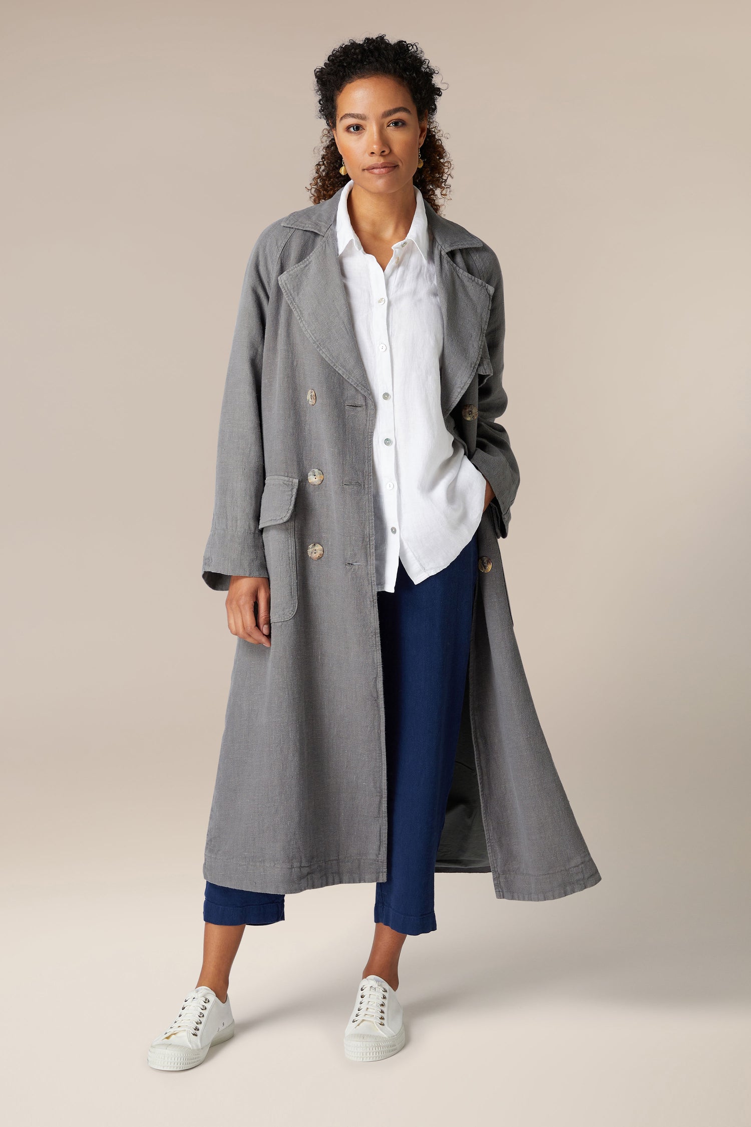 Sentence with Product Name: A woman stands wearing a gray Linen Trench Coat, white shirt, and navy trousers with white sneakers, against a neutral background.