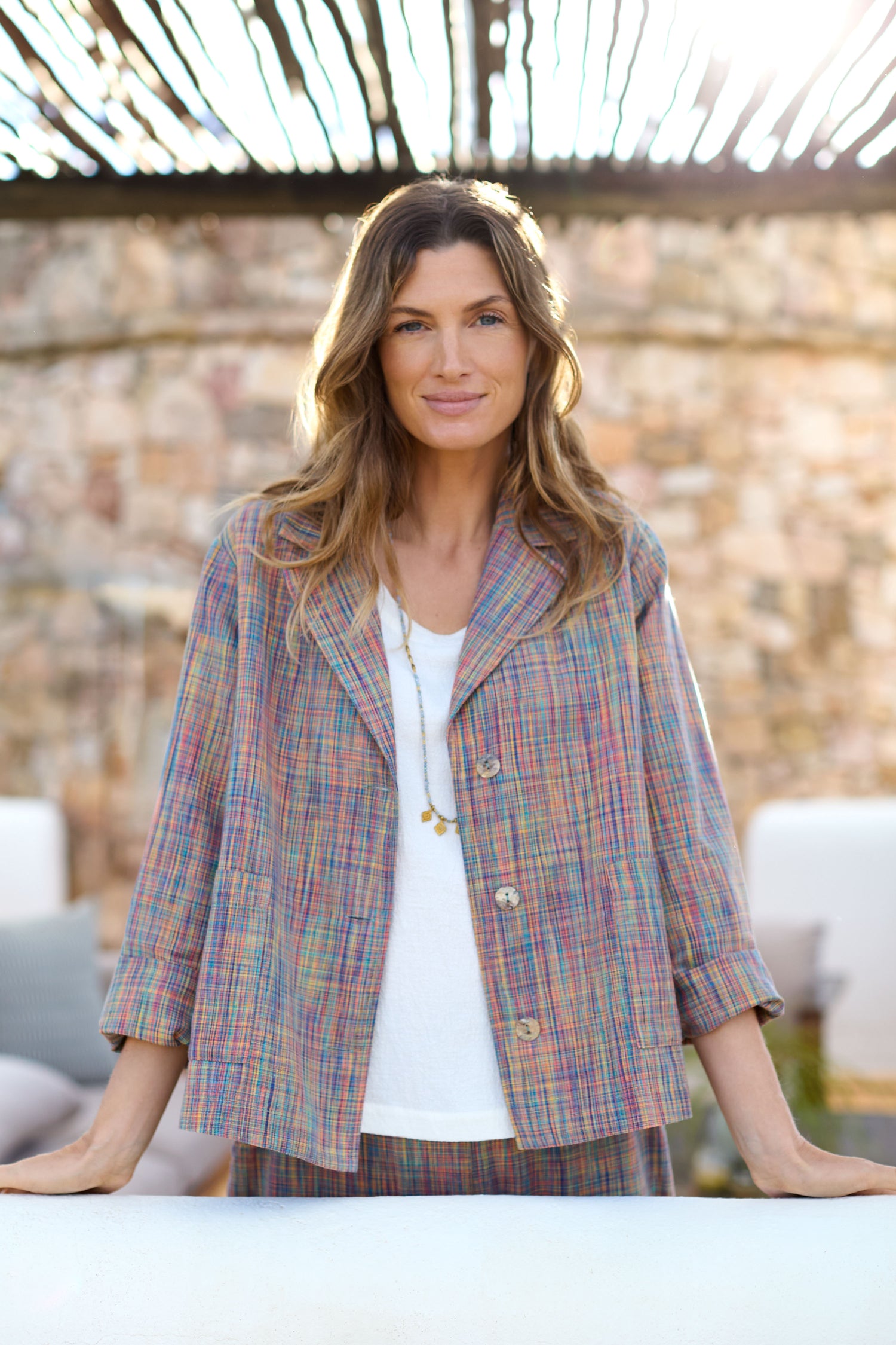 Sporting a Spacedye Crosshatch Jacket over a white shirt, a smiling person stands outdoors with hands on a surface, perfectly matching the stone wall and slatted roof in the background.