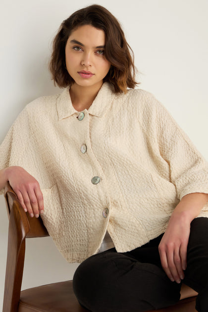 A woman with short brown hair wears the Waffle Linen Boxy Jacket, a cream-colored, boxy button-up, as she sits on a wooden chair against a plain background.