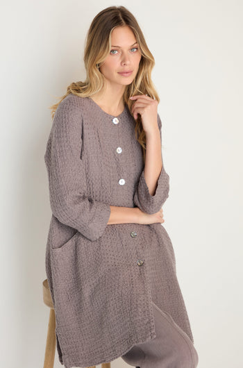 A woman with long blonde hair wears the Waffle Linen Long Jacket and matching pants, true wardrobe essentials, while sitting on a stool against a plain background.