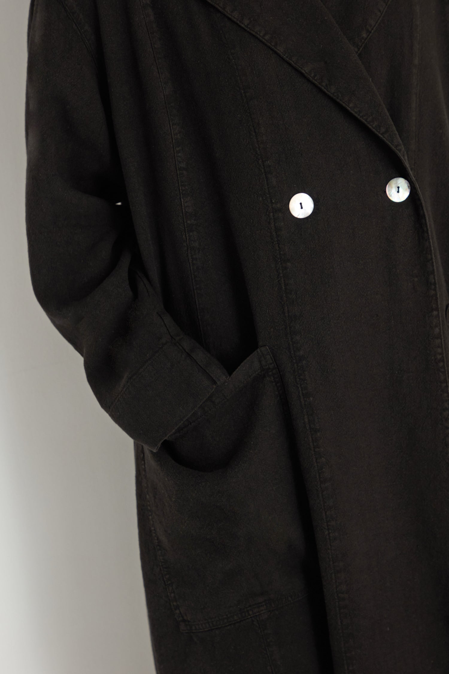 Close-up of a person wearing the Twisted Linen Relaxed Coat in black European linen, featuring two silver buttons and a large front pocket, showcasing a slow fashion approach against a plain background.