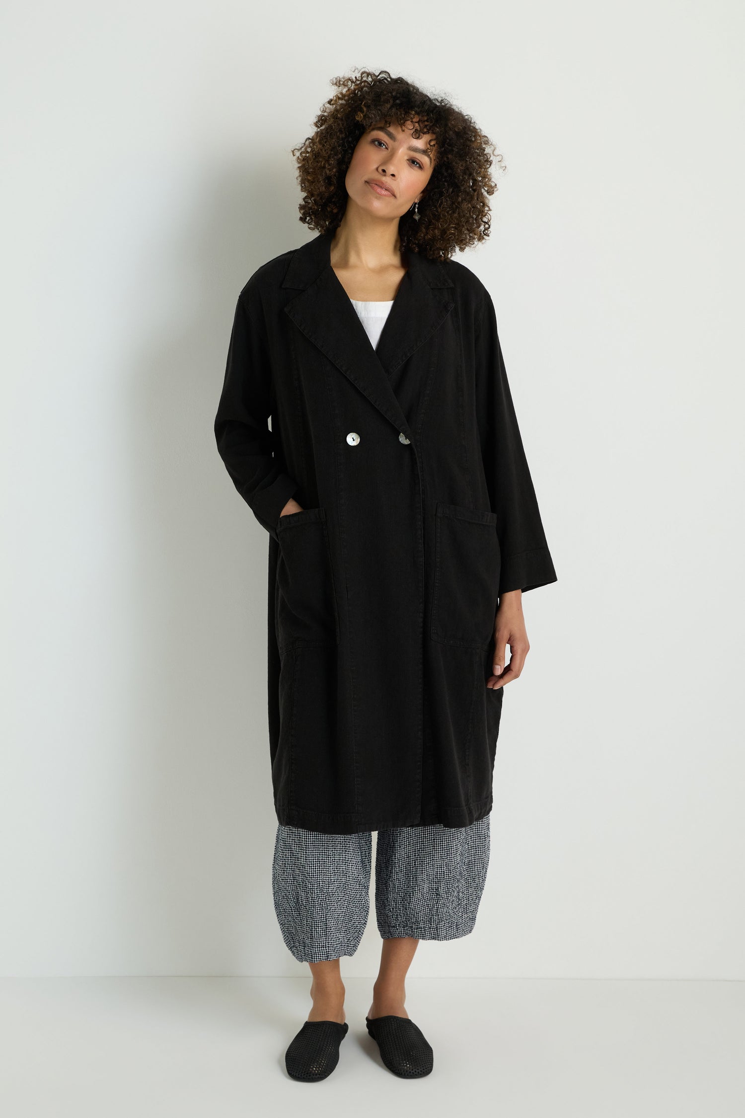A person with long brown hair is wearing the Twisted Linen Relaxed Coat in black over a light top, standing against a plain white background.