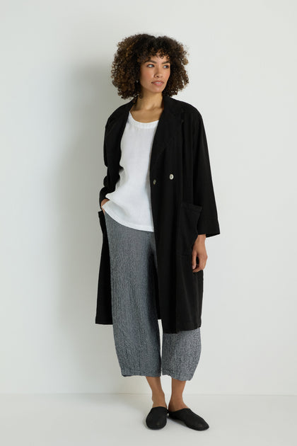 A person stands against a white wall wearing the Twisted Linen Relaxed Coat, a white top, black and white plaid pants, and black sandals—embracing the essence of slow fashion.