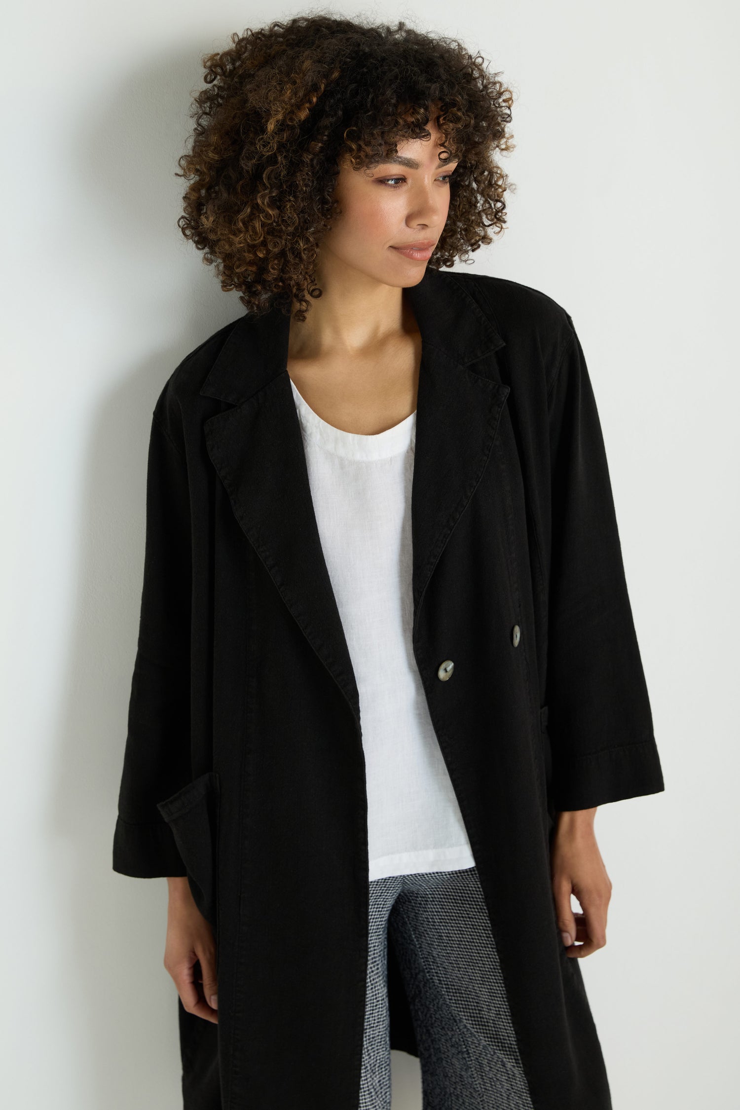 A woman with curly hair wears the Twisted Linen Relaxed Coat over a white top and patterned pants, standing against a plain light-colored wall.