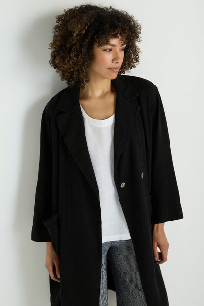 A woman with curly hair wears the Twisted Linen Relaxed Coat over a white top and patterned pants, standing against a plain light-colored wall.