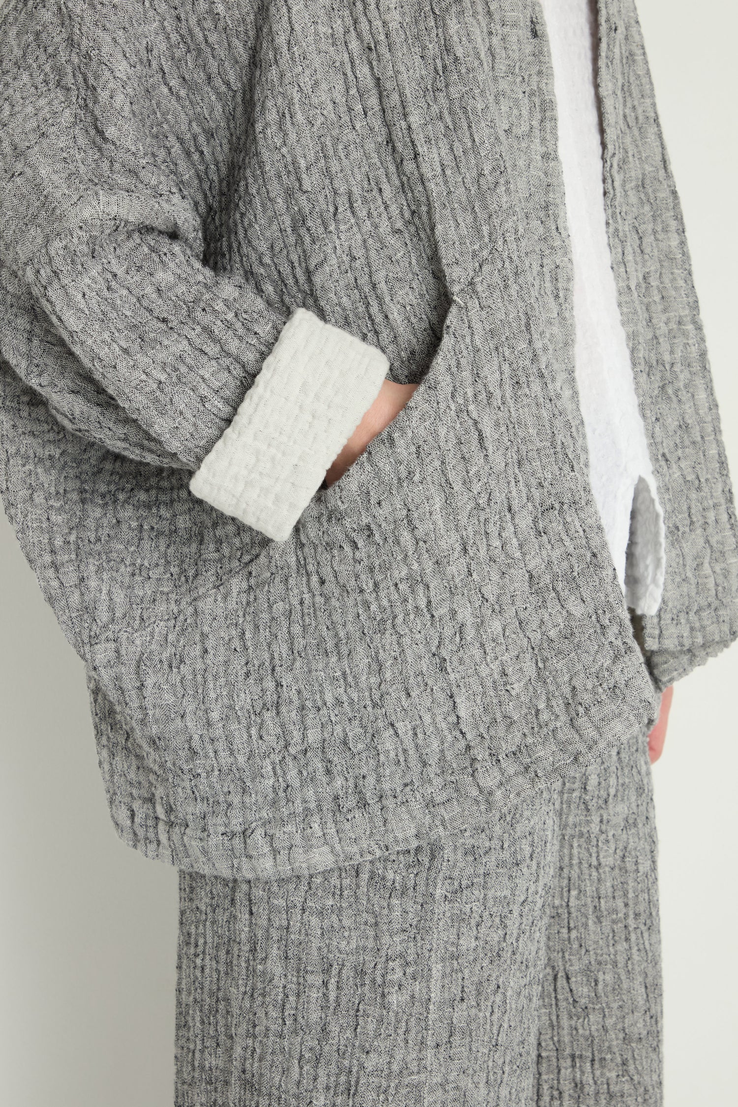 A person stands in front of a plain background wearing the Crinkle Double Face Linen Jacket in textured grey with matching pants, one hand in the pocket.